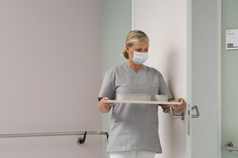 Asistenta medicala cu tava in mana deschide usa cu antebratul, apasand pe manerul anticovid.