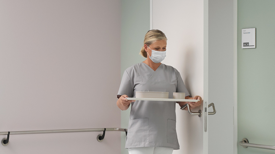 Asistenta medicala cu tava in mana deschide usa cu antebratul, apasand pe manerul anticovid.